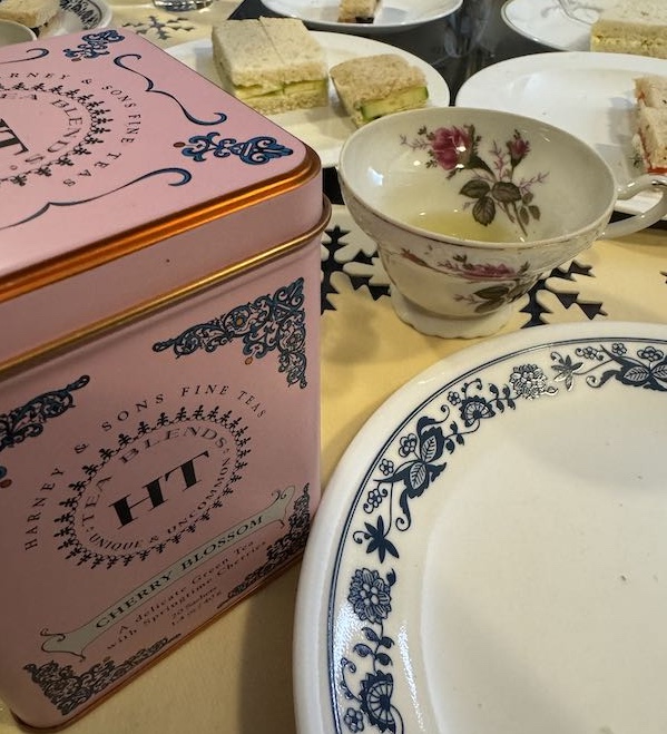 A photo of an at-home Mother's
Day tea with a pink tin of Harney and sons fine teas Cherry Blossom tea, a vintage china tea cup decorated with roses, and tea sandwiches with cucumber, egg salad, and salmon and cream cheese.